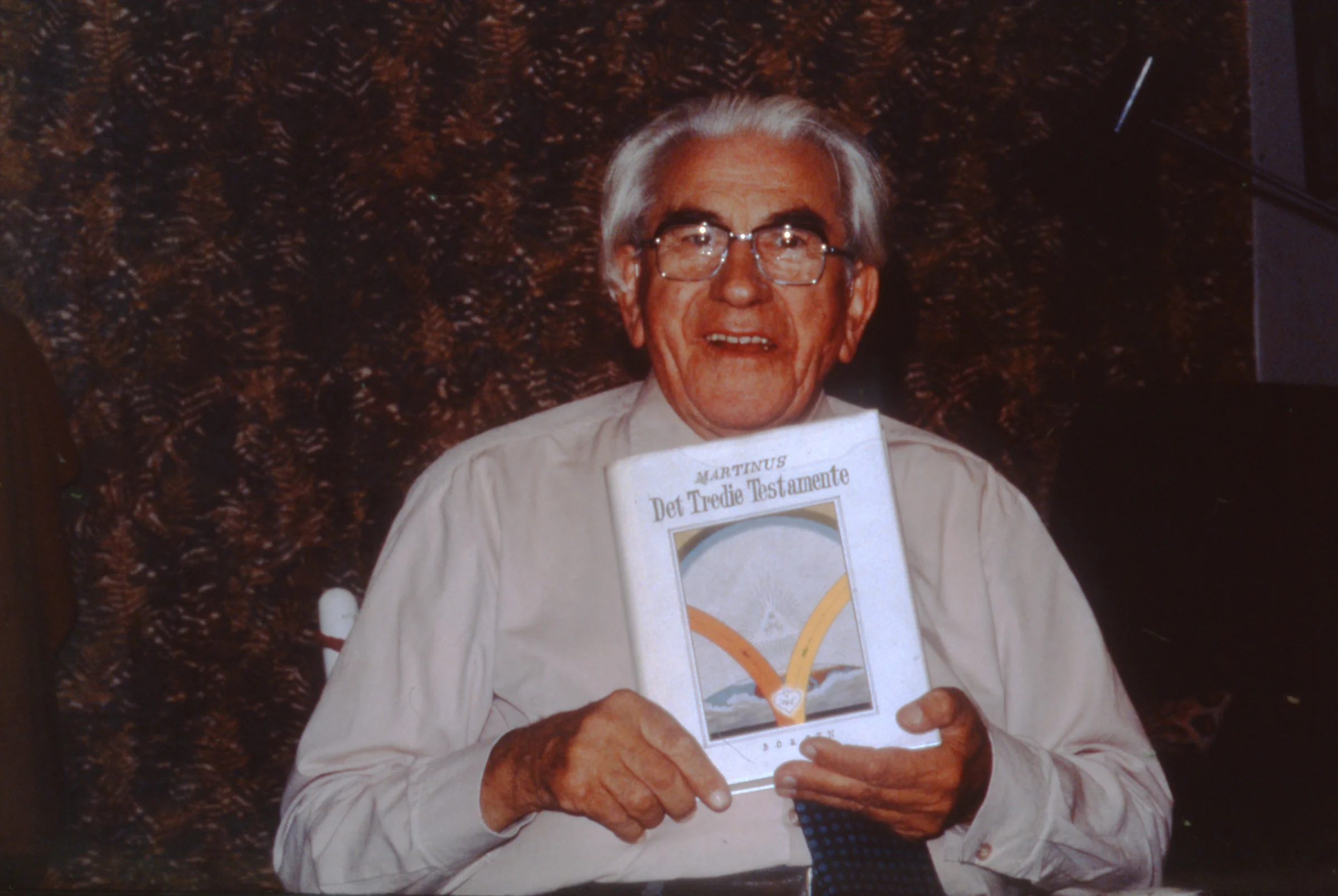 En glad Martinus viser sin layout til præsentationsbogen Det Tredje Testamente. Man bemærker, at der ikke står Livets Bog 1 på forsiden. Det kom først i 1980. Fotografiet er taget 13. august 1978 på Vanløse Alle 26,st.th., hvor jeg genkender stolen og det mørke blomstrede tapet, da jeg selv senere kom til at bebo lejligheden i knapt et års tid. Foto Inge Sørensen., Vanløse Allé 26 st.th. www.martinus.dk